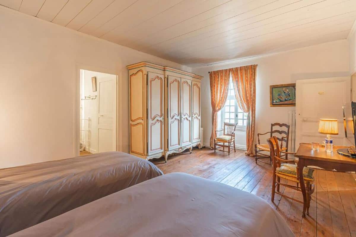 Ein Zimmer mit zwei Betten, Holzboden, einem Esstisch mit Stühlen, einem Schrank und einem Fenster mit Vorhängen.