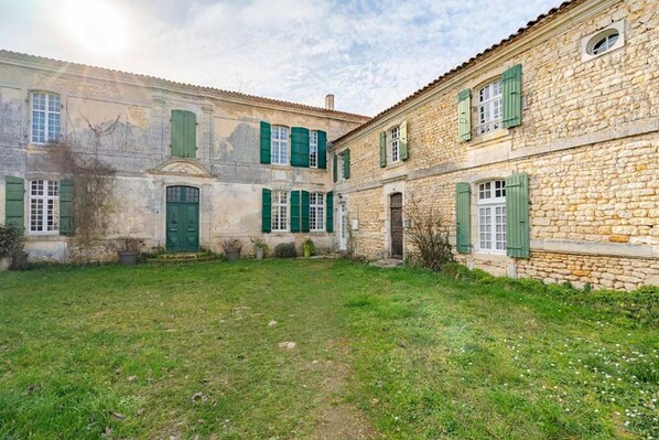 A courtyard with stone buildings, green shutters, and a grassy area. - Maison Garesché (Nieulle-sur-Seudre)
