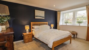 A bedroom with a wooden bed, bedside tables with lamps, a window with curtains, and a painting on the wall. - The Stables (Thornhill)