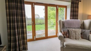 A living room with a sofa, a view through a sliding glass door to a garden, and a wooden ceiling beam. - The Stables (Thornhill)