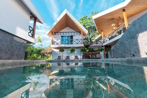 Een modern resort met een zwembad met glazen bodem, een gebouw van twee verdiepingen met een houten dak, en een stenen muur.