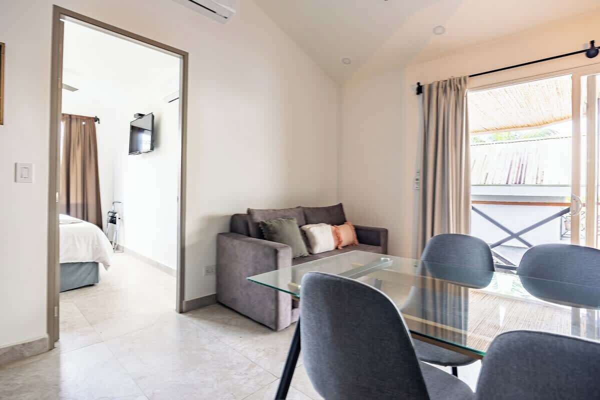 A modern room with a glass table, grey chairs, a sofa with cushions, a TV mounted on the wall, and a sliding glass door leading to an outdoor area.