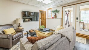 Una sala de estar con sofá, sillón, mesa de centro y televisor. Hay una puerta que da a otra habitación y una ventana con una planta.
