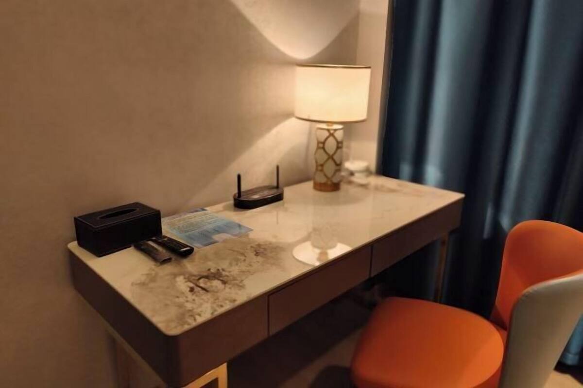 A hotel room with a marble desk, a lamp, a chair, and a small table with a tissue box and remote control.