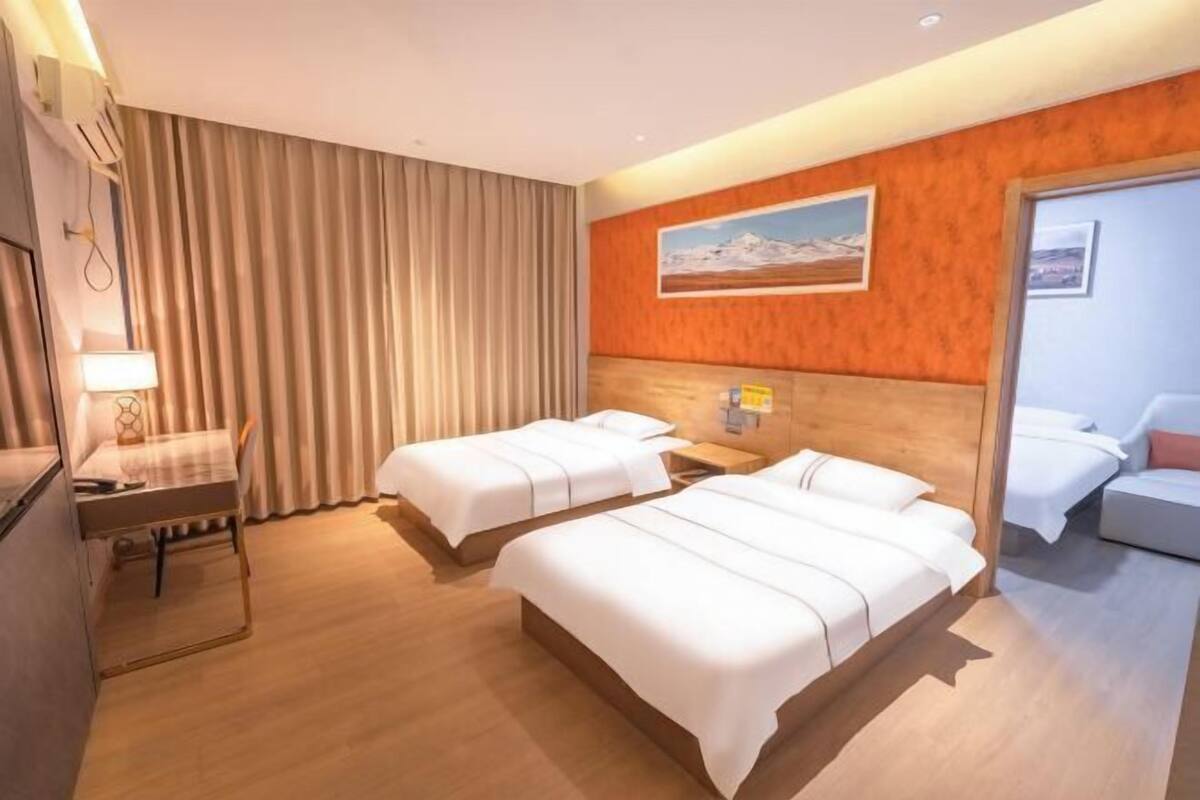 A hotel room with two beds, a desk, a chair, a lamp, a painting on the wall, and a door.