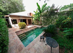 Una piscina con zona de patio, rodeada de abundante vegetación y una casa con tejado de baldosas.