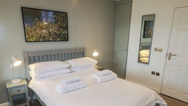 Un lit bien fait, avec du linge de lit et des serviettes blanches, une lampe de chevet et un tableau représentant un paysage naturel accroché au mur.