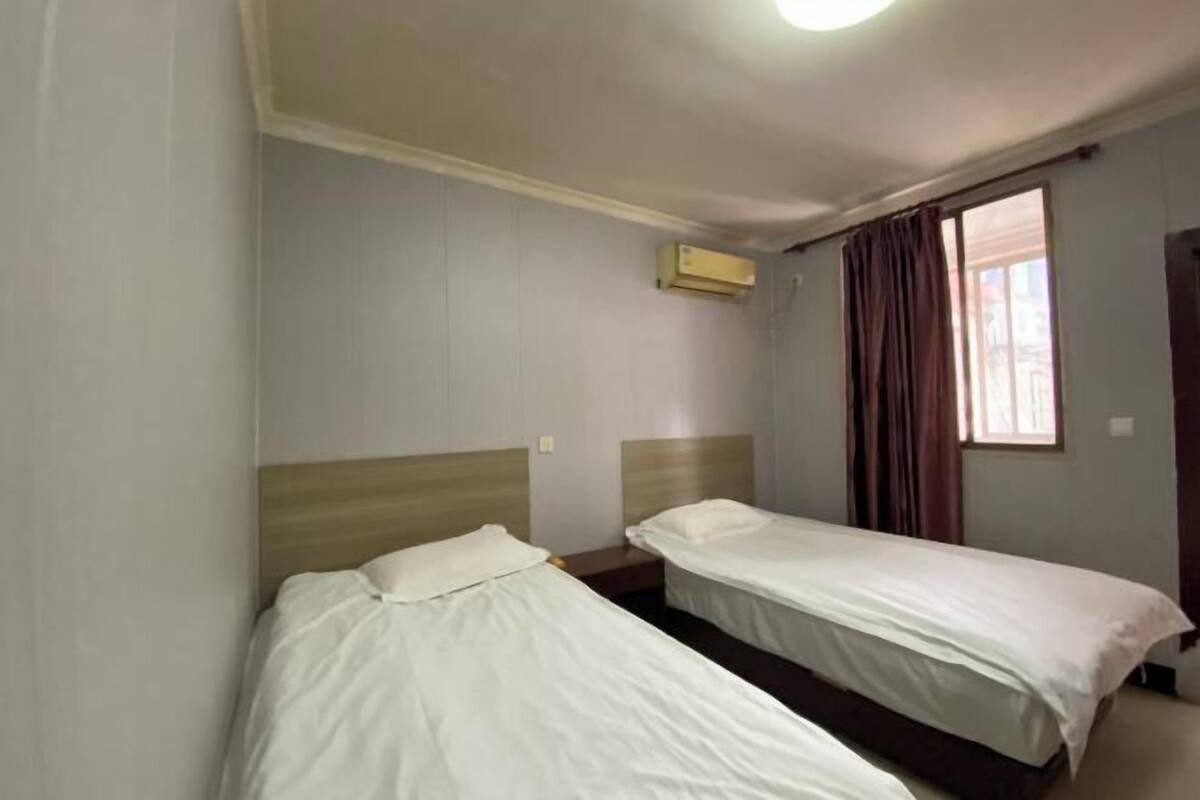 A hotel room with two single beds, a window with curtains, an air conditioning unit, and a wooden headboard.