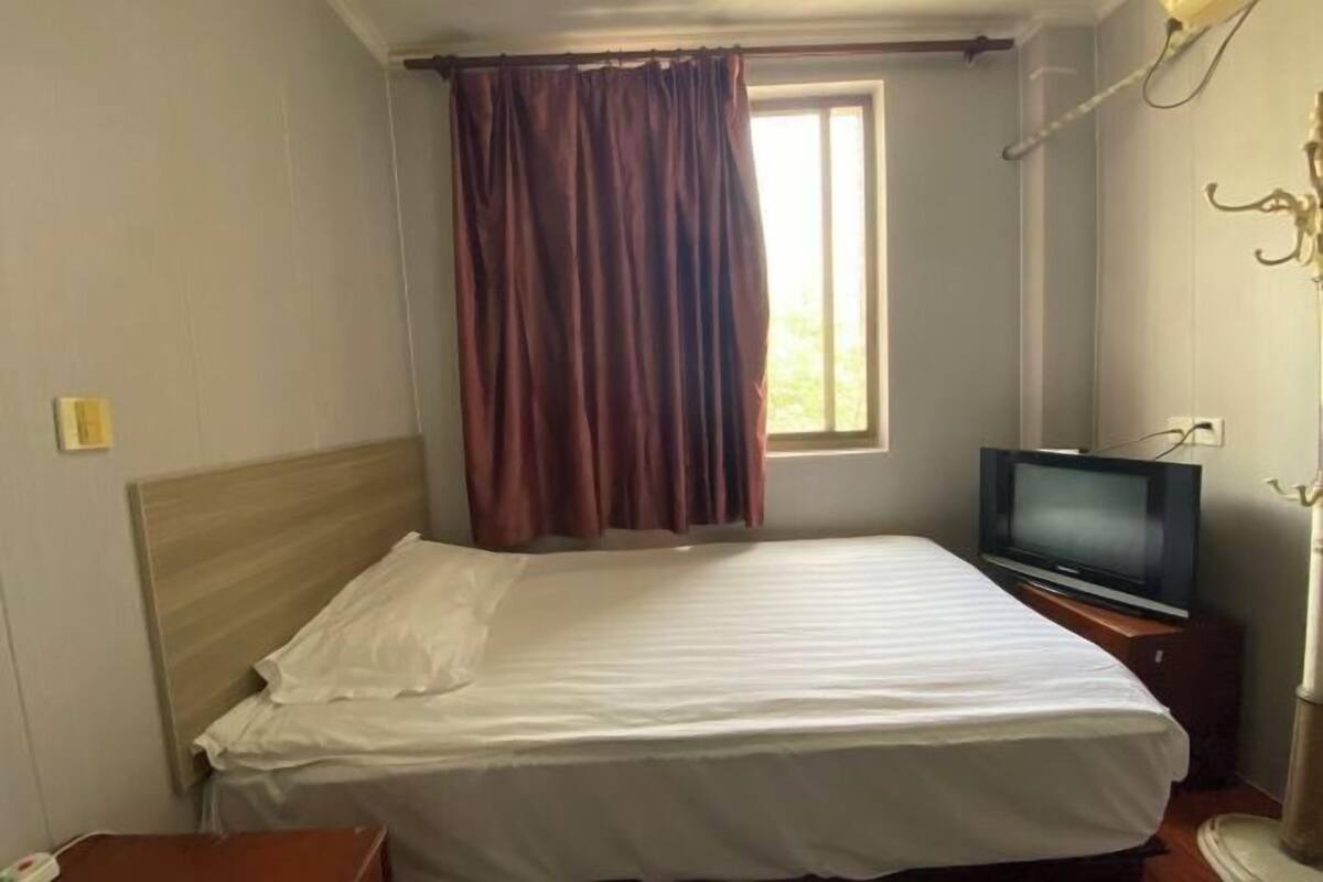 A hotel room with a bed, a television, a window with curtains, and a bedside table.