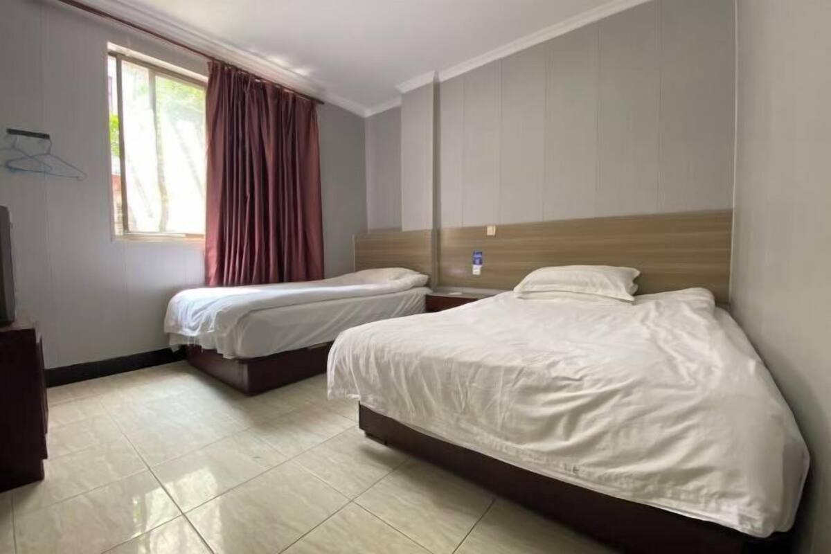 A hotel room with two beds, a window with curtains, a wooden headboard, and a tiled floor.