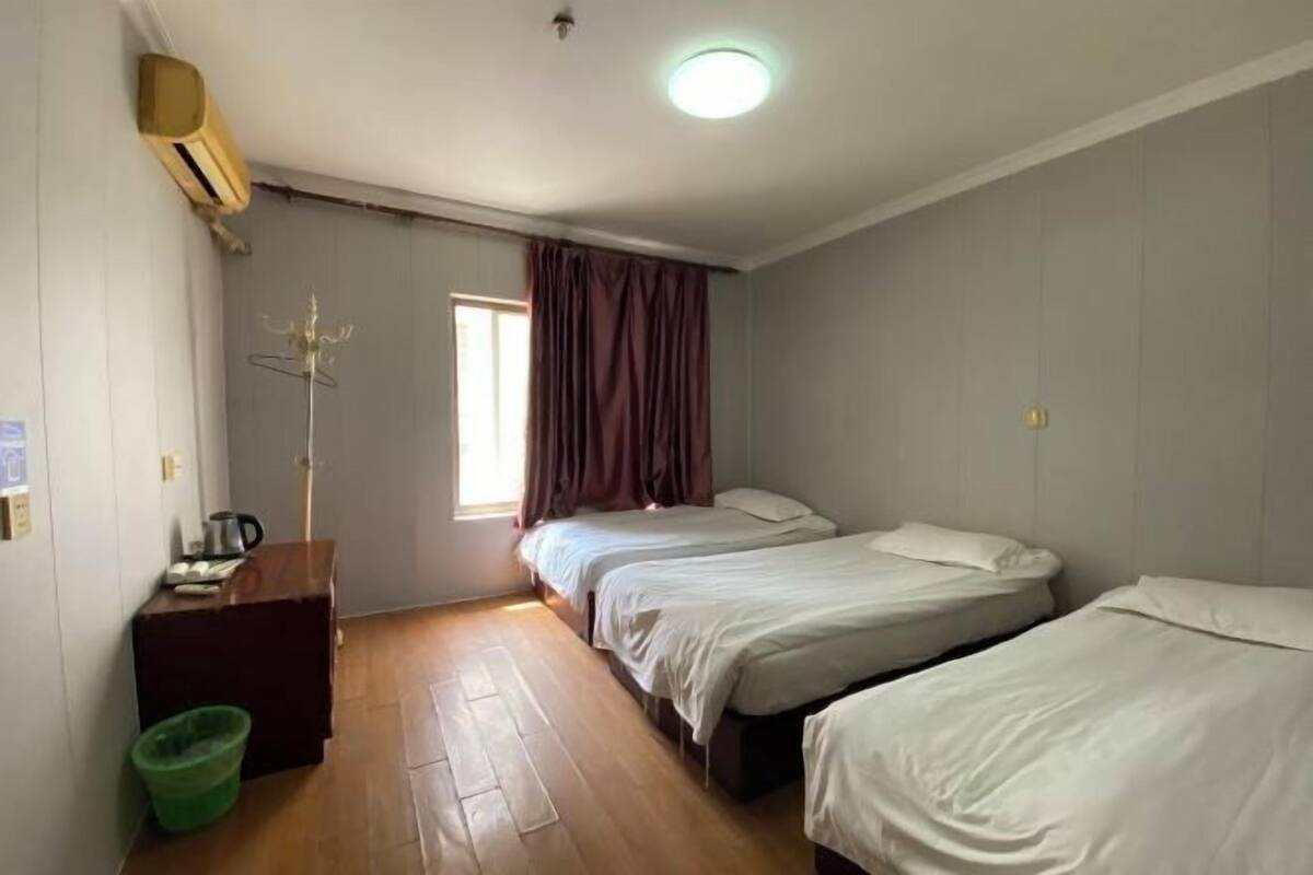 A room with three single beds, a wooden floor, a desk with a chair, a green trash bin, and a window with curtains.