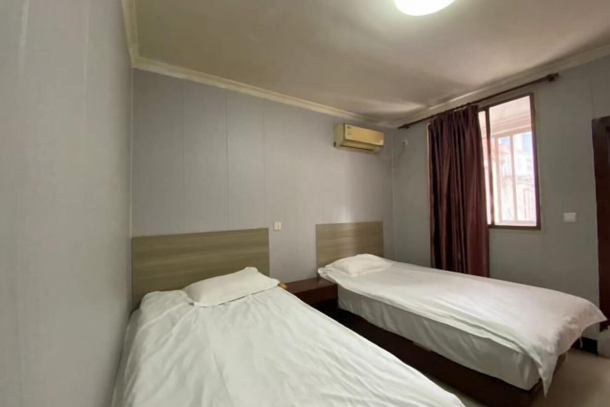 A hotel room with two single beds, a window with curtains, an air conditioning unit, and a light fixture on the ceiling.