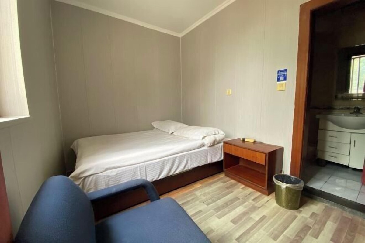 A small, neatly arranged bedroom with a single bed, a nightstand, a chair, and a visible bathroom with a sink and mirror.