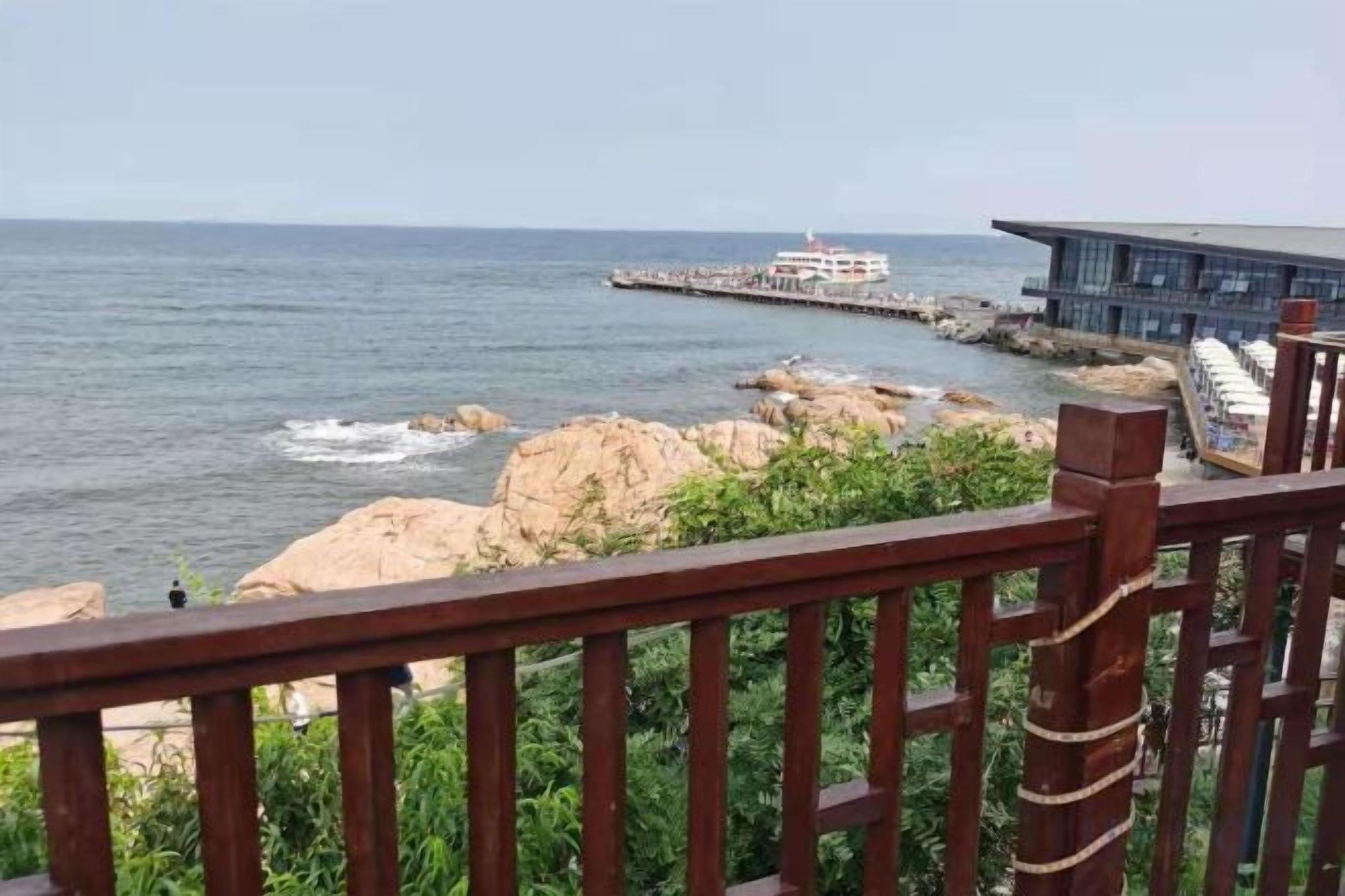 Vista costera con muelle que se adentra al mar, un edificio de diseño moderno y una baranda de madera al frente.