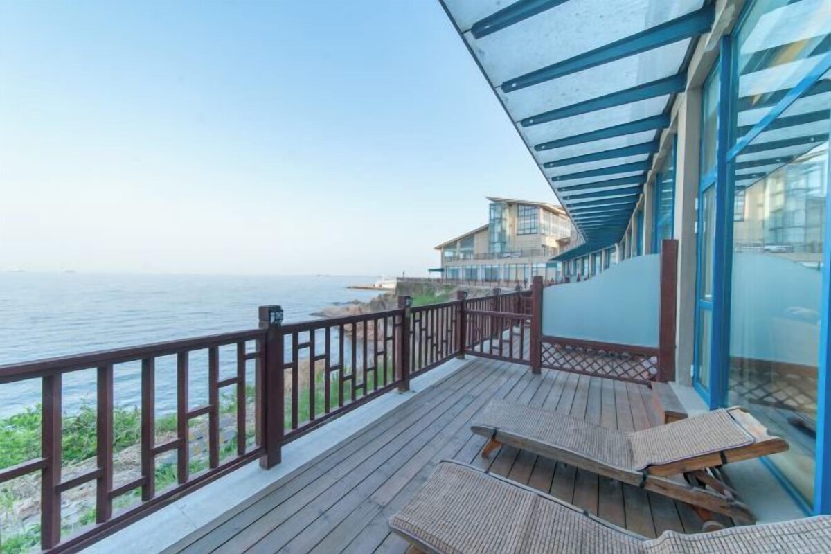 A wooden deck with a glass railing overlooking the sea, a bench, and a building with a blue roof.