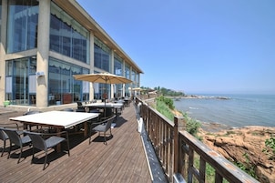 Una terraza de madera con mobiliario exterior, con vista a un cuerpo de agua y un litoral rocoso.