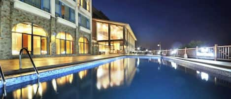 Ein Schwimmbecken mit Holzterrasse und einem Gebäude mit vielen Fenstern und einer Steinfassade.