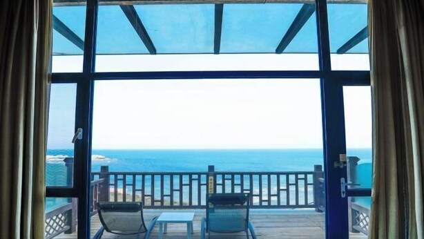 A room with a large window overlooking the ocean, two chairs, a small table, and a wooden deck. - Beidaihe Bei Hua Yuan Sea View Hotel (Beidaihe Biluo Tower) (Qinhuangdao)