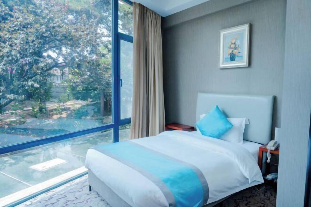 Una camera d'albergo con una grande finestra, un letto con biancheria da letto bianca e blu, un comodino con un telefono e un quadro appeso al muro.