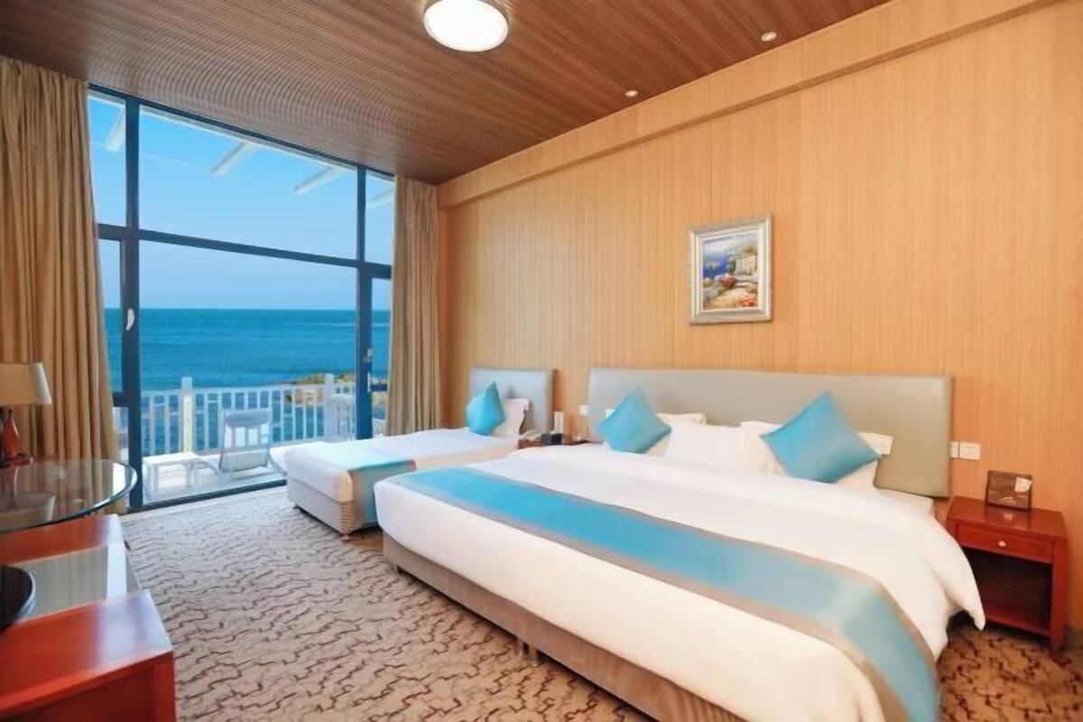 Camera d'albergo con due letti, un'ampia finestra vista mare, un piccolo balcone e un soffitto in legno.