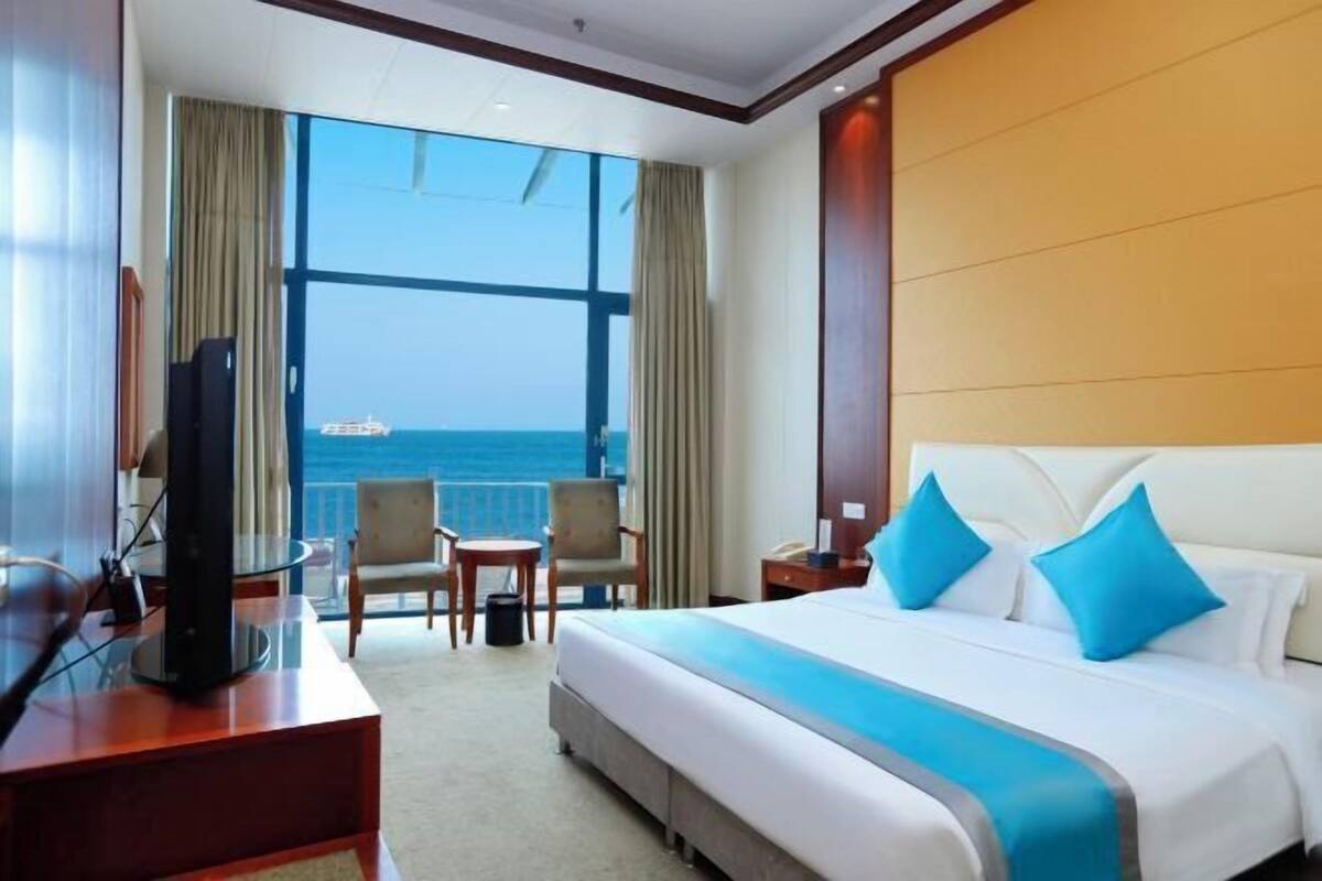 Una camera d'albergo con un'ampia finestra, un letto con cuscini blu, una scrivania con televisore e una sedia.