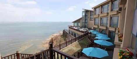 A hotel balcony with a view of the sea, outdoor seating with umbrellas, and a staircase leading to the upper floors.