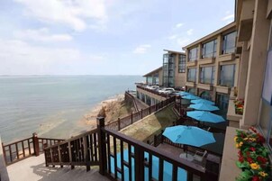 Balcón de hotel con vista al mar, mobiliario exterior con sombrillas y una escalera que conduce a los pisos superiores.