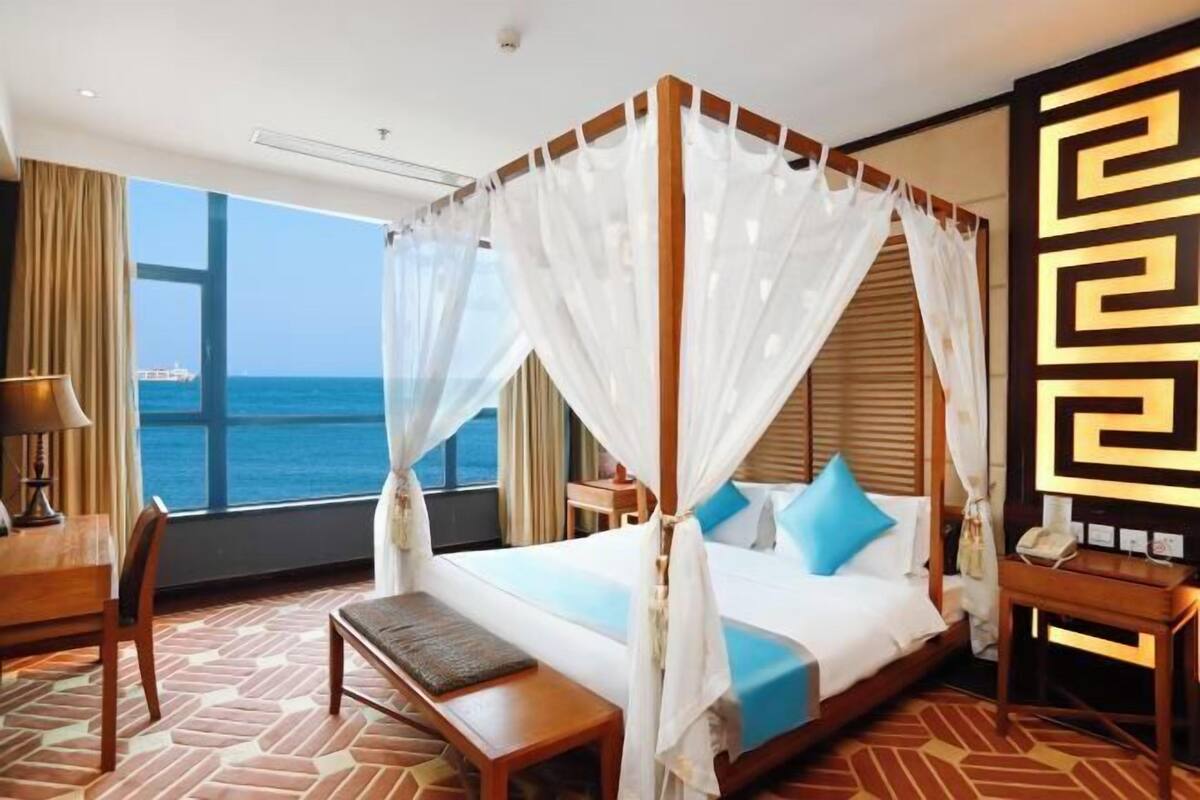 Un letto a baldacchino con tende leggere, una panca in legno, una scrivania con una lampada e vista sul mare.