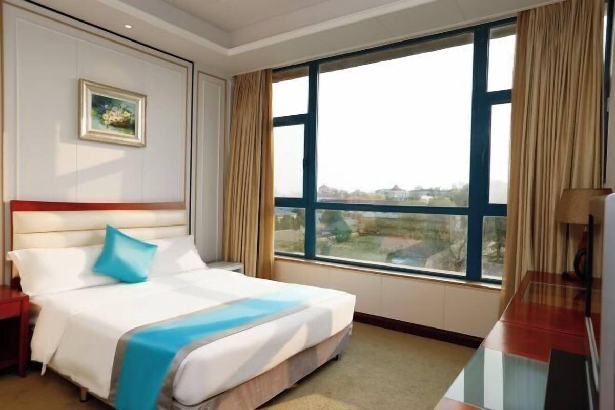 Una camera d'albergo con una grande finestra, un letto con biancheria bianca e un dettaglio blu, un quadro appeso al muro e un comodino in legno.