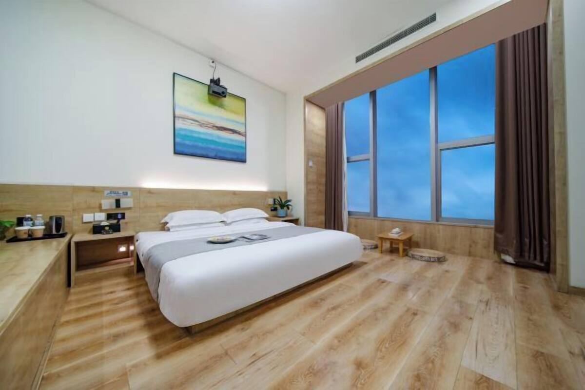 A modern hotel room with a large bed, a nightstand, a small table, and a large window offering a view of the sky.