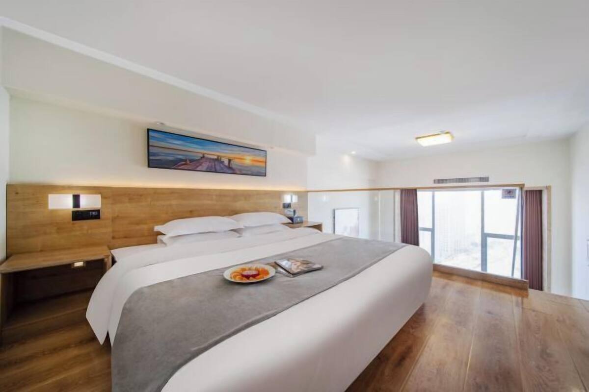 A modern hotel room with a large bed, wooden headboard, bedside tables, and a view through a glass door.