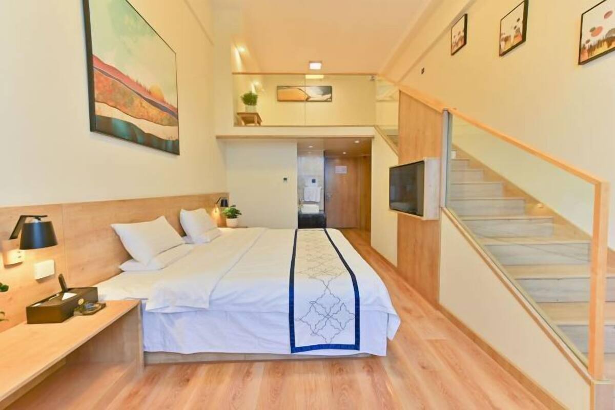 A hotel room with a bed, a desk with a lamp, a TV, a staircase, and a painting on the wall.