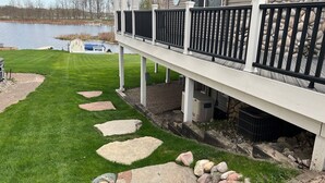 A house with a stone wall, a deck with black railings, and a stone pathway leading to the house. - Lakefront Getaway on Murray Lake - Swim, Fish, Float, & Relax (Lowell)