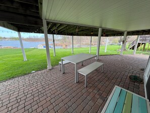 Eine überdachte Terrasse mit Tisch und Bank, Backsteinböden und Blick auf einen See.