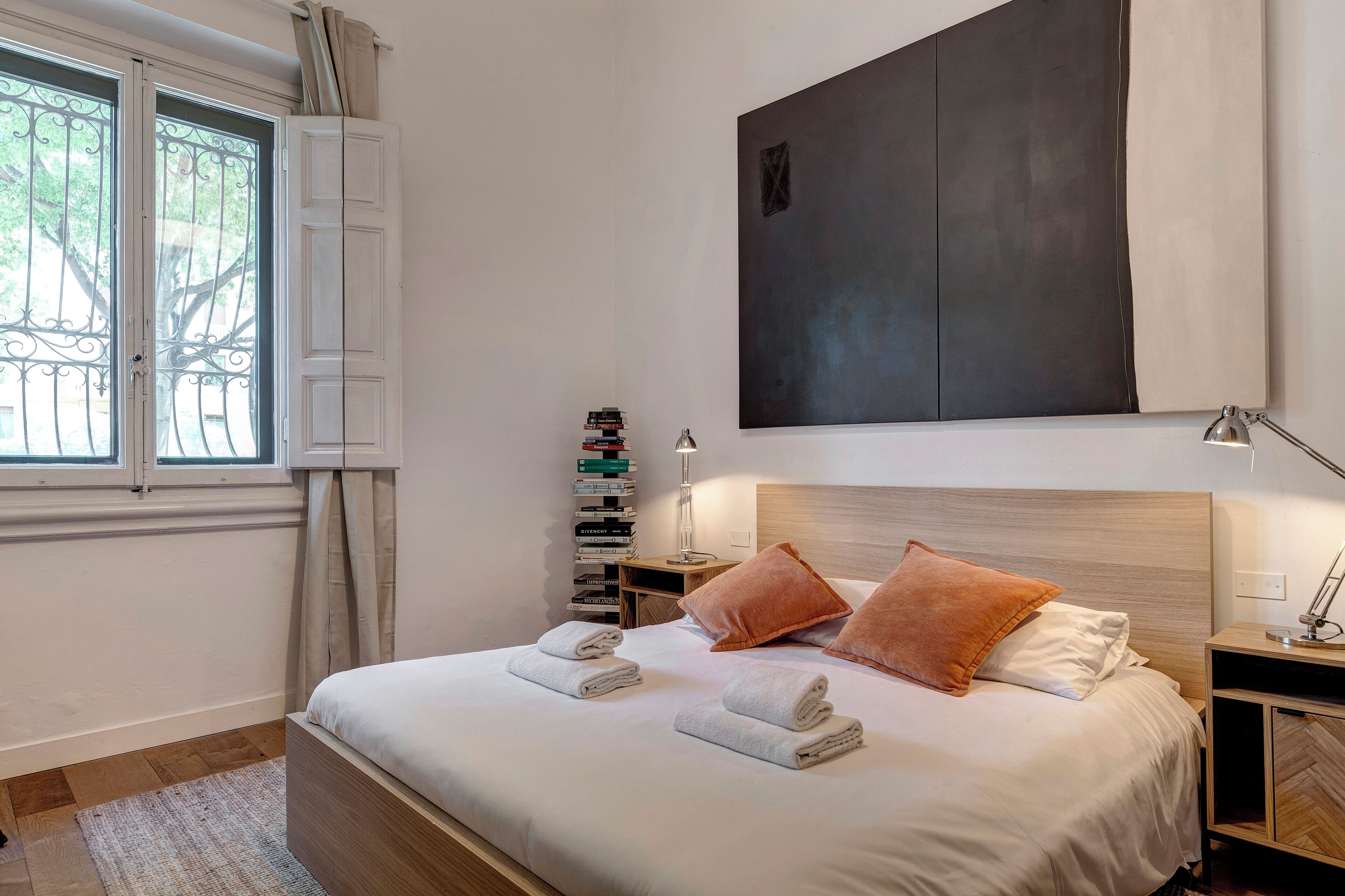 A neatly made bed with towels, a bedside table with a lamp, a stack of books, and a large abstract painting on the wall.