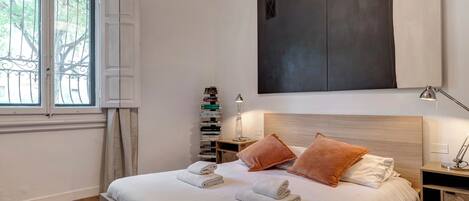 A neatly made bed with towels, a bedside table with a lamp, a stack of books, and a large abstract painting on the wall.
