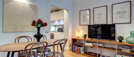 A dining area with a wooden table and chairs, a vase with red roses, and a bookshelf with books and decorative items.