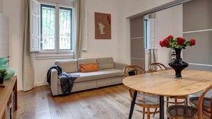 En stue med sofa, spisebord med stole, et vindue med gardiner, en potteplante og en vase med blomster.