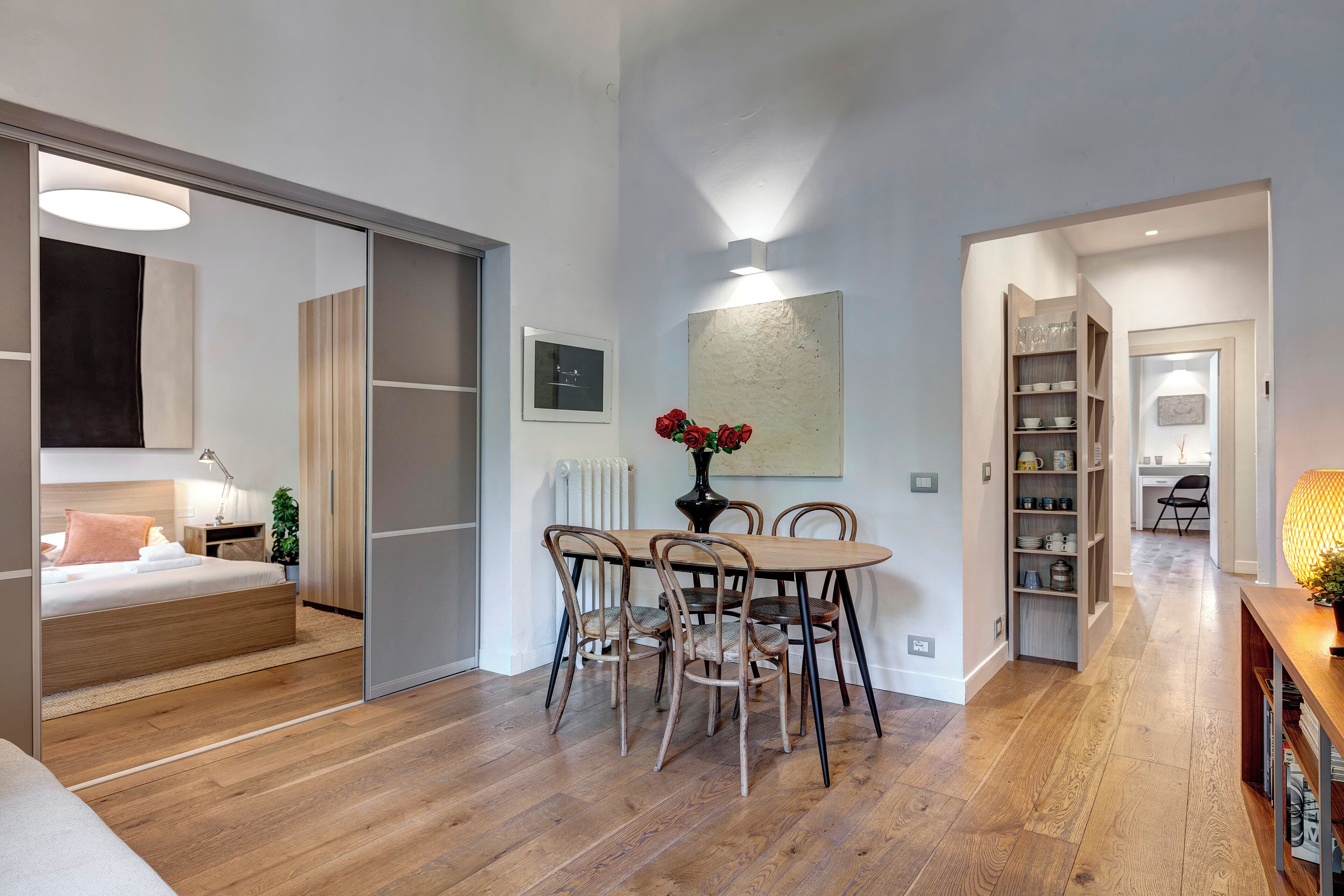 A modern room with a wooden floor, a dining table with chairs, a bed with a headboard, a radiator, and a bookshelf.