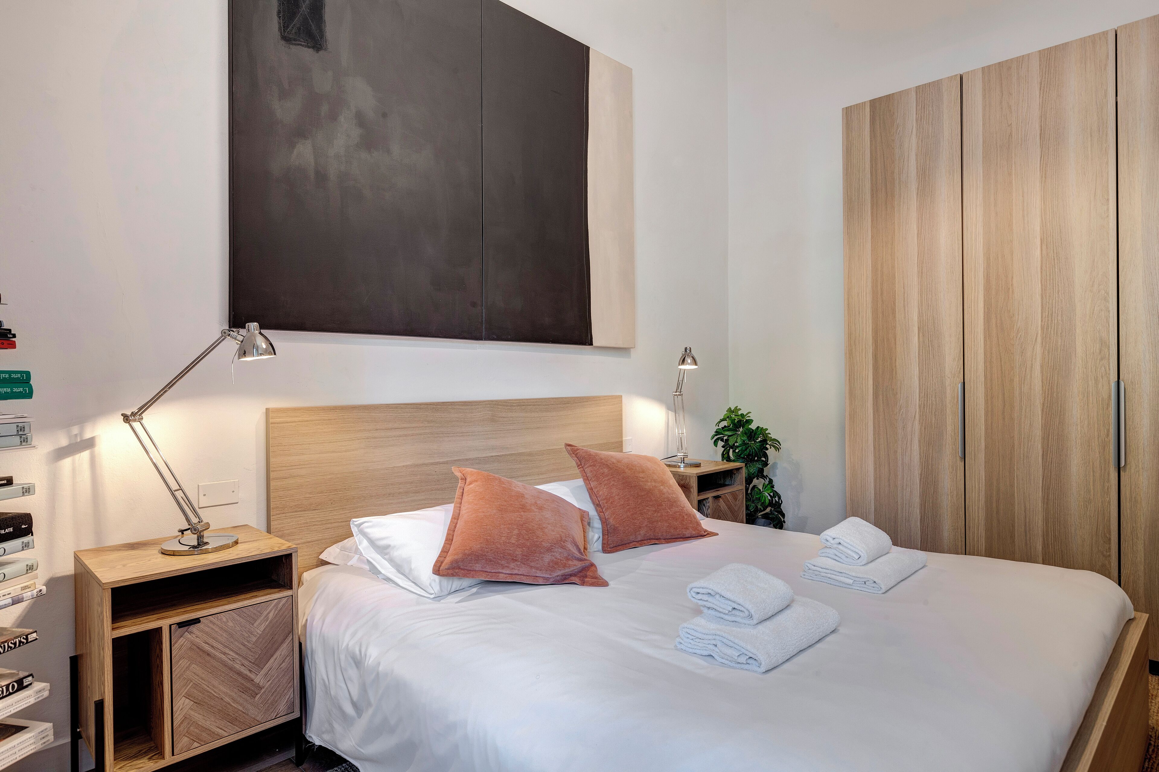 Une chambre d’hôtel moderne avec un grand lit, deux oreillers, une table de chevet avec une lampe, une armoire en bois et un tableau abstrait accroché au mur.