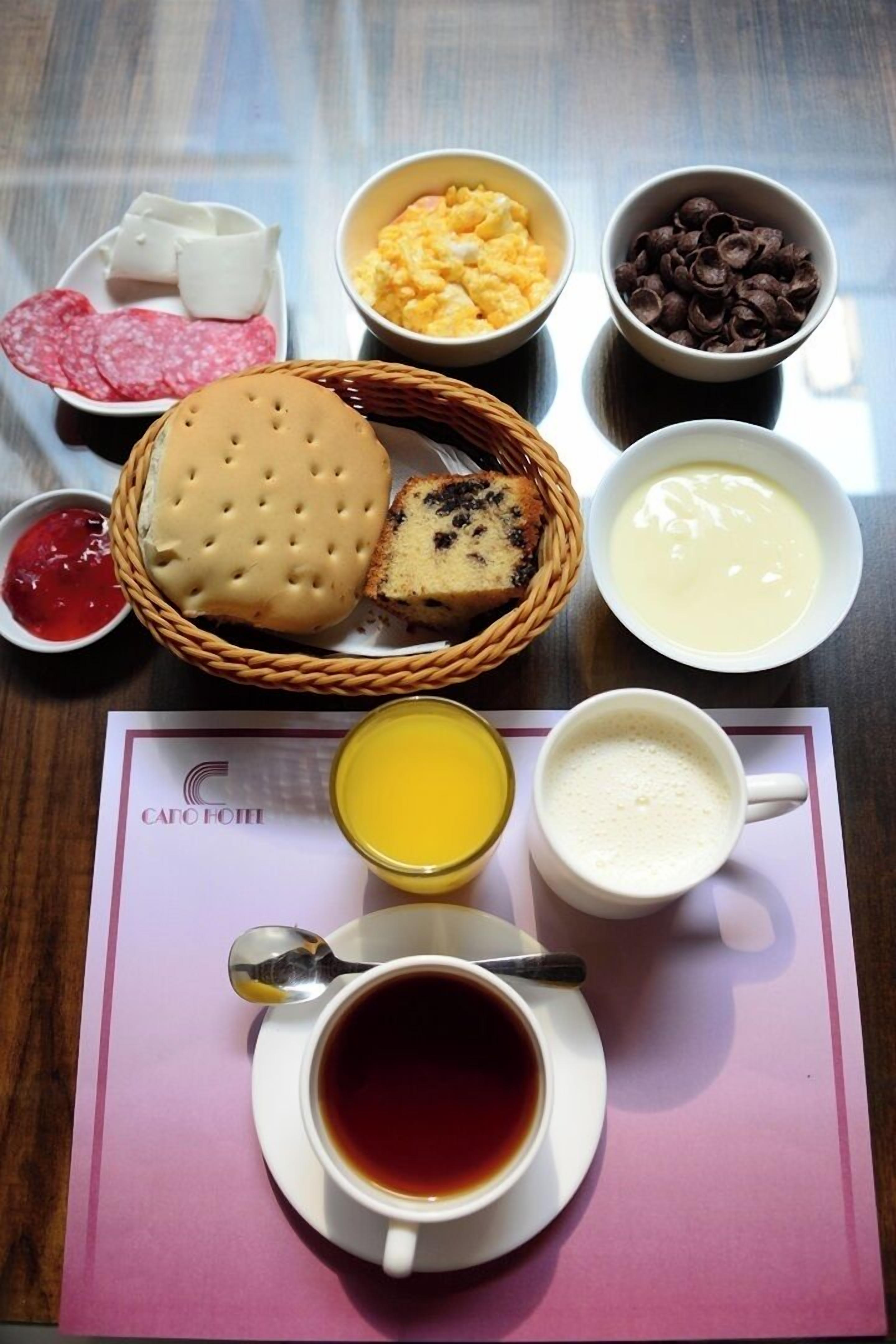 A breakfast setting with a basket of bread, a plate of salami, scrambled eggs, chocolate chips, and a cup of coffee.