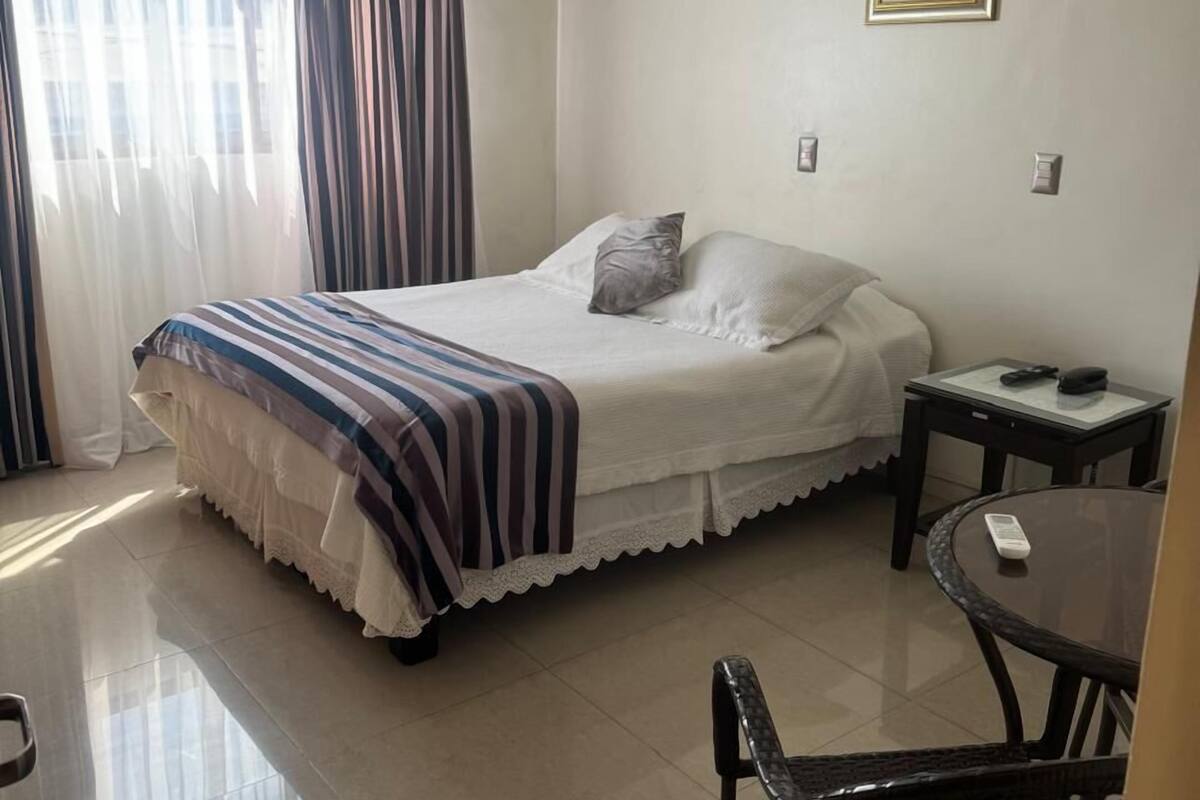 A hotel room with a bed, a small table, a chair, and a window with curtains.
