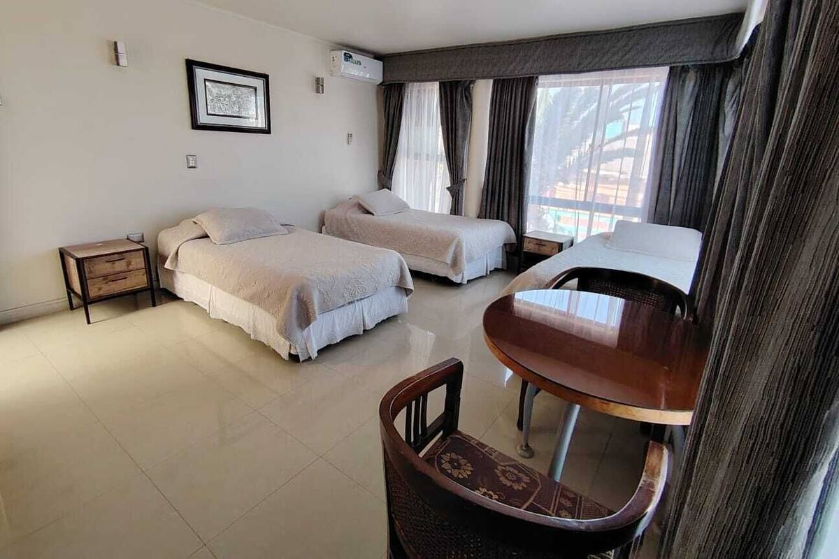 A hotel room with two beds, a small table, a chair, a nightstand, and a window with curtains.