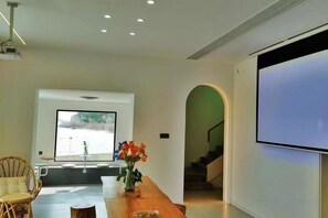 Un salon moderne avec une grande fenêtre, une table basse en bois, un fauteuil en osier et un écran de projection.