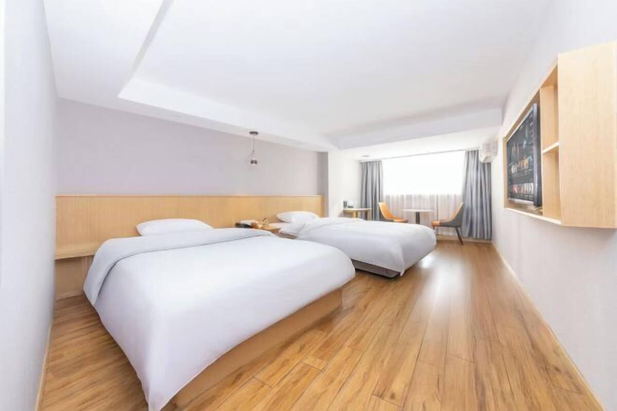 A hotel room with two beds, wooden flooring, a large window with curtains, and a wall-mounted TV.