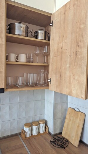 Kitchen area with a shelf holding a pot, kettle, mugs, and pitcher, along with a cutting board and tumblers.