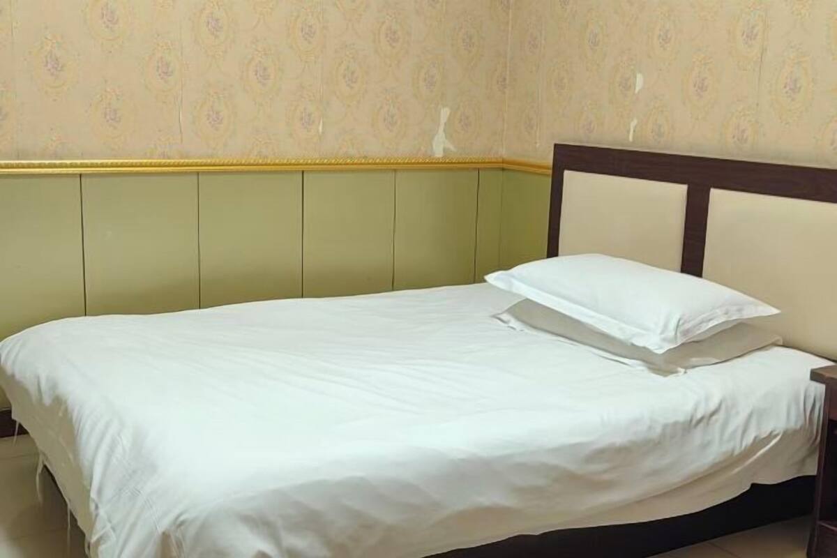 A single bed with a wooden headboard, a green padded headboard, and a plain wall with a floral pattern.