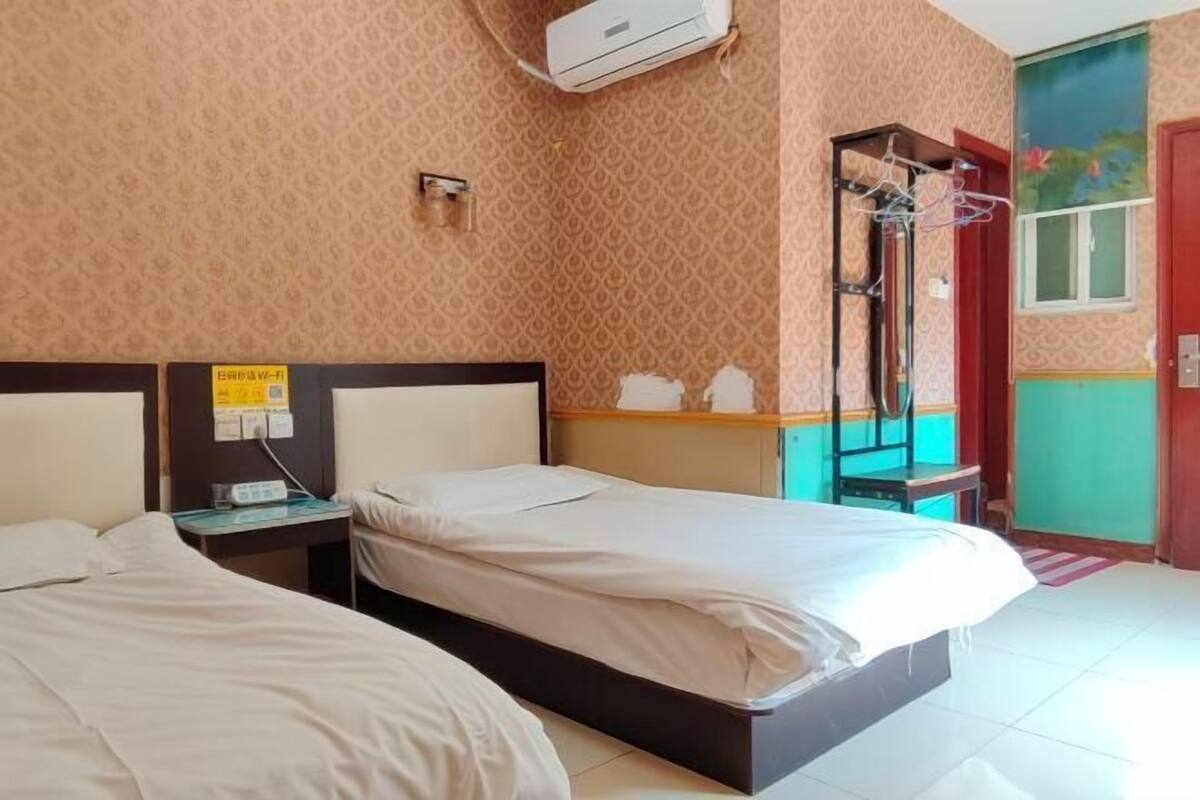 A hotel room with two beds, a bedside table, an air conditioning unit, a wall-mounted heater, and a window with a view.