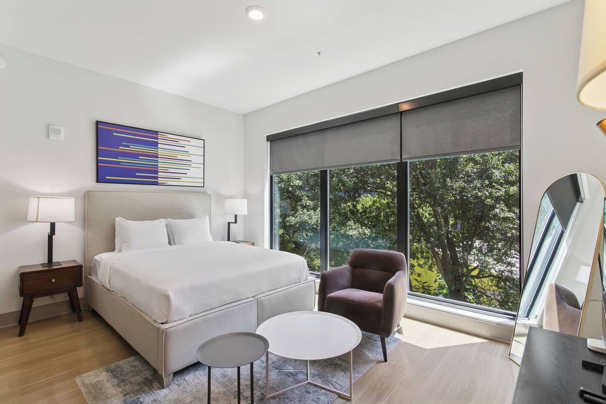 A modern bedroom with a large bed, a side table, a chair, a small table, and a large window with a view of trees.