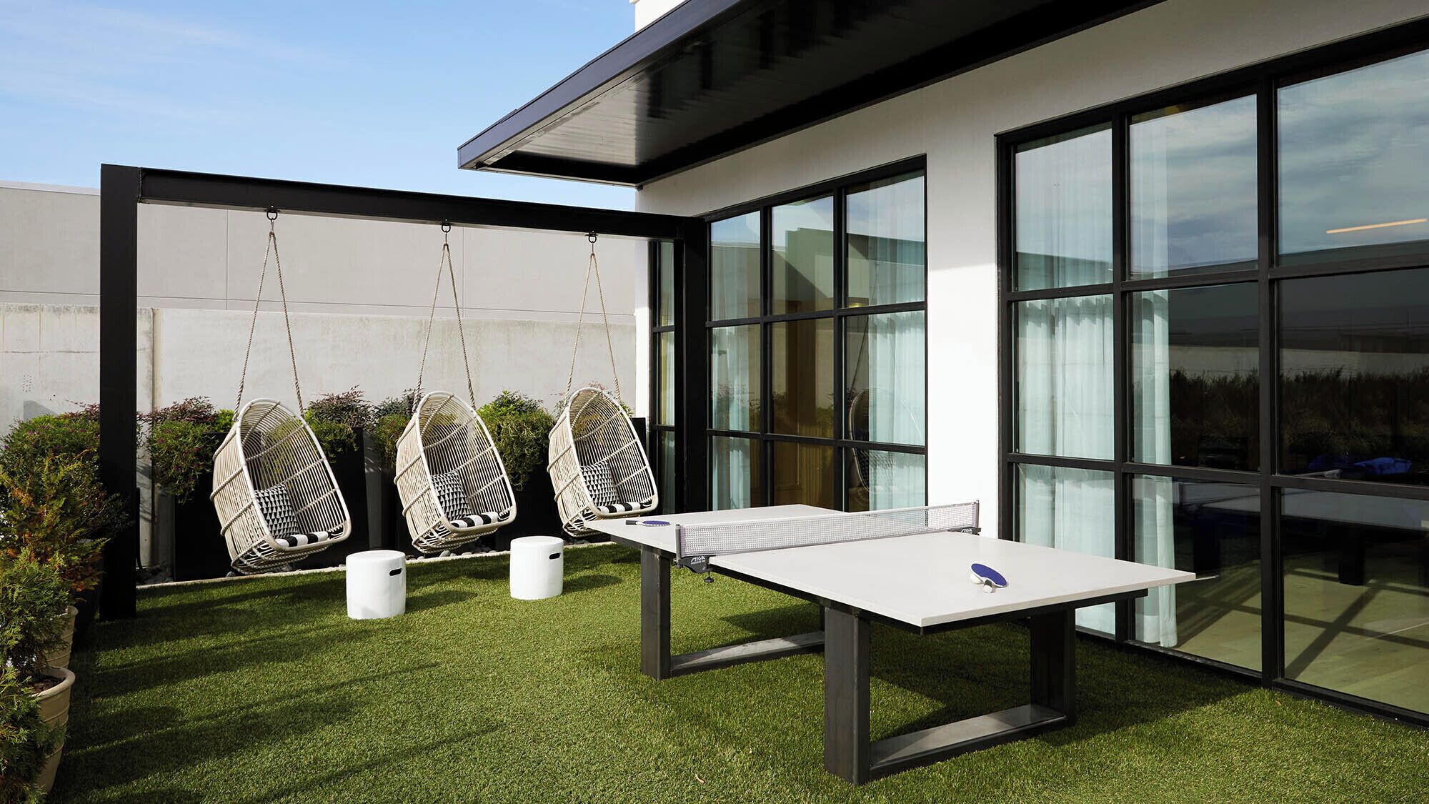 A modern outdoor space with a ping pong table, three hanging chairs, and a glass-enclosed area with large windows.